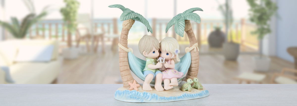 A Precious Memories figurine of a couple sitting in a hammock between two palm trees, displayed on a light-colored surface with a blurred indoor background.